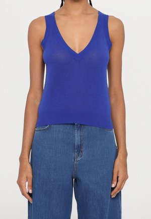 Femme portant un haut en maille bleu sans manches à col en V associé à un jean en denim bleu ample à taille haute, montré du cou aux hanches.