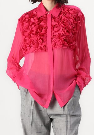 Jasnoróżowa przezroczysta bluzka z trójwymiarowymi kwiatami z tkaniny na piersi, długimi rękawami i kołnierzykiem, zestawiona z szarymi spodnimi w kratę.