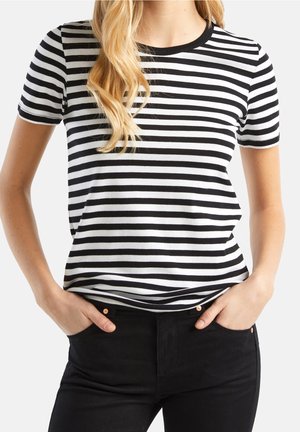 Kvinde med langt blondt hår iført en sort og hvid t-shirt med vandrette striber og sorte bukser, hænderne i lommerne.