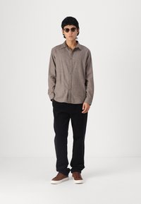 Καφέ πουκαμίσα με σχέδια και μακριά μανίκια, μαύρο παντελόνι καστόρι και καφέ αθλητικά παπούτσια με λευκές σόλες. Το μοντέλο φοράει σκούρα γυαλιά ηλίου και στέκεται μπροστά.