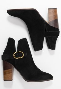 Botines de ante negro con una silueta curvada, acento de hebilla circular dorada y un tacón de bloque de madera a rayas. Textura mate, sin patrones visibles.