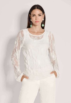 Vrouw met lang donker haar, gekleed in een wit bloemig kanten top met lange mouwen en witte broek, met mintgroene en gouden hangende oorbellen.