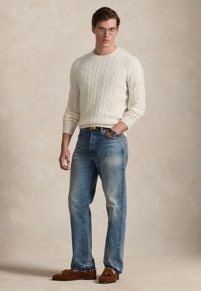 Camisola de malha torcido com gola redonda, cor marfim; jeans azul claro com bainha desfiada; mocassins castanhos. Modelo está em pé com as mãos nos bolsos.