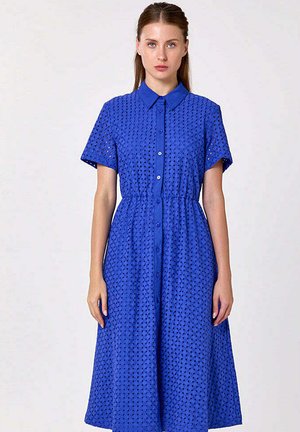 Femme debout portant une robe bleu roi à œillets, longueur genou, avec manches courtes, col et boutons devant, sur un fond uni.