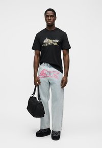 Μαύρο μπλουζάκι με πολυχρωματικό γραφικό, ανοιχτό μπλε τζιν παντελόνι με ροζ περίγραμμα καρτούν χαρακτήρα και μαύρη τσάντα.