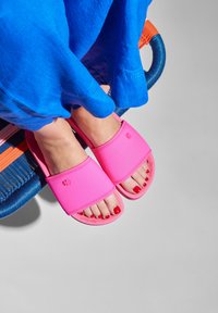 Pink rubber slide sandals with a flat sole and stitched detailing, paired with a blue fabric garment and red toenails visible.