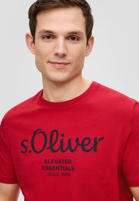 Rode katoenen T-shirt met een zwart bedrukte logo en de tekst "s.Oliver ELEVATED ESSENTIALS SINCE 1969." Ronde halslijn en korte mouwen.