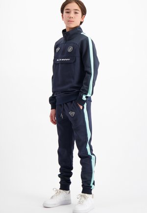 Marineblauwe trainingspak met mintgroene zijstrepen, een met rits afsluitbare zak op de borst, geribbelde boorden en logodetail op de broek. Witte sneakers maken de outfit compleet.