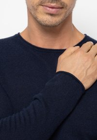 Navy-blauer Strickpullover mit Rundhalsausschnitt, gerippten Bündchen und weicher Textur. Die Hand des Modells ruht sanft auf dem Schulterbereich.