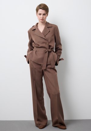 Persoon met kort haar, gekleed in een bruine blazer met ceintuur en bijpassende wijde broek, staand tegen een effen witte muur.