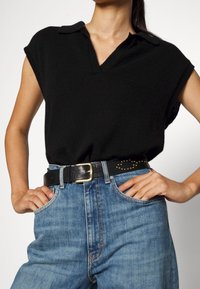 Schwarzes ärmelloses Oberteil mit Kragen, kombiniert mit hoch taillierten blauen Jeans und einem schwarzen Gürtel mit goldenen Nieten.