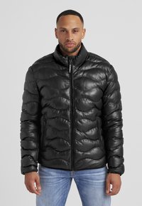 Schwarze Pufferjacke mit glänzender, gesteppten Textur und hohem Kragen. Mit Reißverschluss vorne und seitlichen Taschen, kombiniert mit hellblauen Jeans.