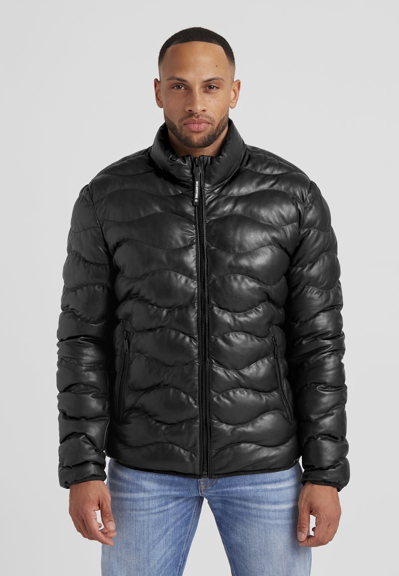 Schwarze Pufferjacke mit glänzender, gesteppten Textur und hohem Kragen. Mit Reißverschluss vorne und seitlichen Taschen, kombiniert mit hellblauen Jeans.