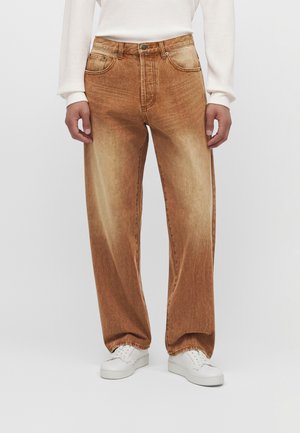 Personne portant un jean large marron délavé, un haut blanc à manches longues et des baskets blanches, debout devant un fond clair uni.