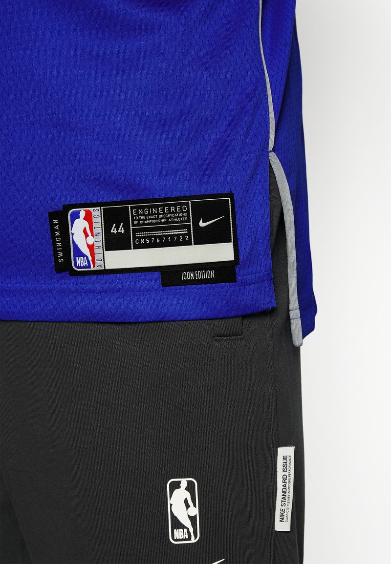 Royal niebieska koszulka z siateczkową teksturą, z czarną etykietą Nike i brandingiem NBA. Czarne spodenki poniżej z logo oraz metkami w odcieniach szarości i bieli.