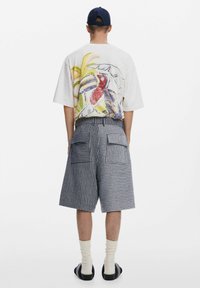 Kék és fehér csíkos nadrág, melyen két hátsó zseb található. A pólón egy színes tropikus madár látható világos háttérrel. Fekete szandál van rajta.