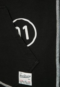 Camiseta negra con el número "11" impreso en blanco y logotipo. Presenta una etiqueta con el nombre de la marca "Raw Denim" y las instrucciones de cuidado. Costuras laterales visibles.