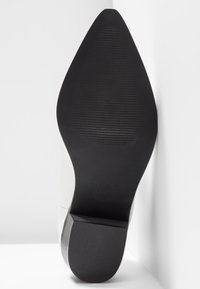 Semelle noire pointue d'une chaussure féminine avec des lignes de grip texturées et un talon carré, taille 37, inclinée contre une surface blanche.