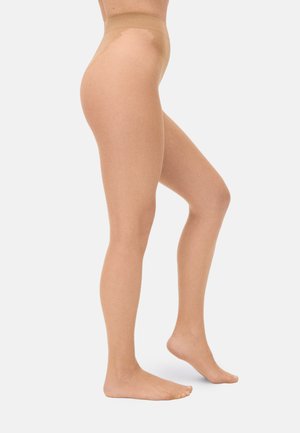 Mujer con medias transparentes color nude, de pie sobre un pie con el otro ligeramente levantado, contra un fondo liso.