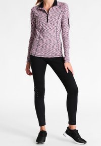 Langärmliges Viertel-Reißverschluss-Oberteil mit einem pinken, grauen und schwarzen Zickzack-Muster; kombiniert mit schwarzen Leggings; verfügt über eine Reißverschlusstasche am Ärmel.