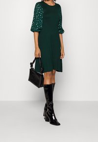 Groene jurk met opstaande mouwen en gouden stippen. Gecombineerd met glanzende zwarte hoge laarzen en een gestructureerde zwarte handtas.