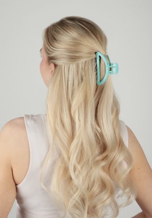 Femme blonde aux longs cheveux ondulés attachés à moitié avec une grande pince à cheveux bleu clair, portant un haut blanc sans manches.
