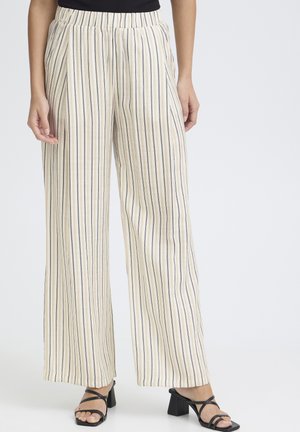 Femme portant un pantalon crème taille haute à jambes larges avec de fines rayures verticales noires et beiges, associé à des sandales noires à talon bloc ouvert à l'orteil.