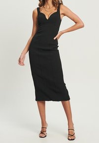 Femme portant une robe midi noire ajustée sans manches avec des fines bretelles et des sandales à talons hauts noires à lanières, se tenant debout avec une main sur la hanche.