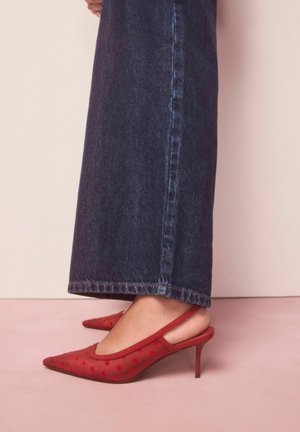 Kāja ar sarkanām tīkliņa stilā slingback augstpapēžu kurpēm ar smalkiem sirds rakstiem, kopā ar tumši ziliem, platajiem džinsiem.