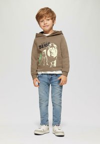 Brauner Hoodie mit Löwen-Graphic und Text, übereinander getragen mit einem weißen Shirt. Hellblaue Jeans. Graue und weiße Turnschuhe. Kurzes, welliges blondes Haar.