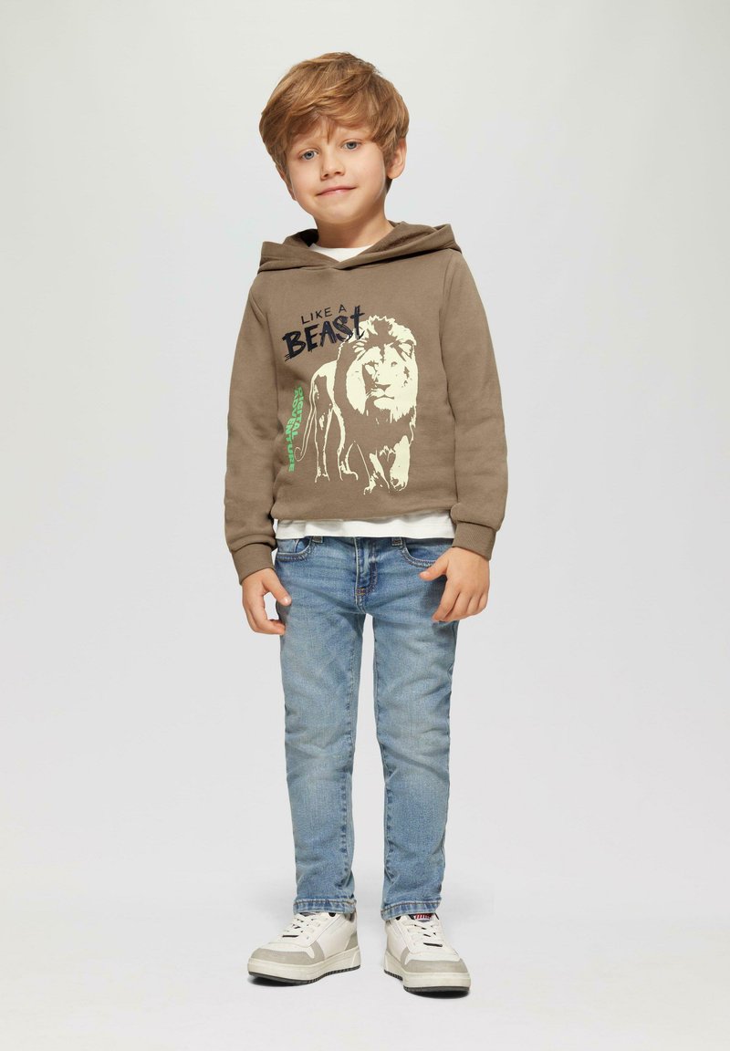Brauner Hoodie mit Löwen-Graphic und Text, übereinander getragen mit einem weißen Shirt. Hellblaue Jeans. Graue und weiße Turnschuhe. Kurzes, welliges blondes Haar.
