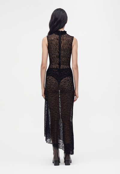 Femme aux longs cheveux foncés se tenant de dos, portant une robe noire transparente sans manches à motif zèbre et des bottes noires.