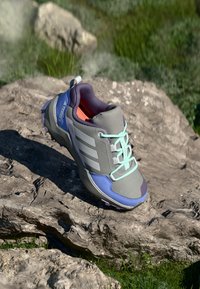 Um único sapato de caminhada cinza, roxo e azul com atacadores azul claro repousando sobre uma superfície rochosa, com erva verde ao fundo.