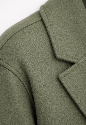 Gros plan sur l'épaule et le large revers cranté d'un manteau en laine vert olive, mettant en valeur la texture du tissu et les détails de la couture.