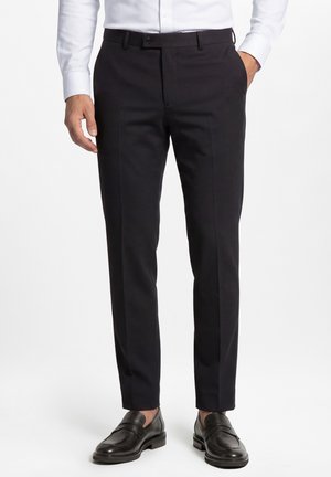 Mann trägt schwarze Slim-Fit-Hose, weißes Hemd und schwarze Lederloafers, steht mit den Händen in der Nähe der Taschen vor weißem Hintergrund.