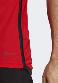 Camiseta deportiva roja con un tejido texturizado, ribete negro a los lados y logo "AEROREADY". El diseño es entallado y de mangas cortas.