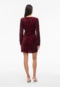 Burgundská mini šaty so zdobením, dlhými rukávmi, z textúrovaného zamatového materiálu, okrúhlym výstrihom a priliehavým pásom. Viditeľné strieborné sandále na vysokom opätku.