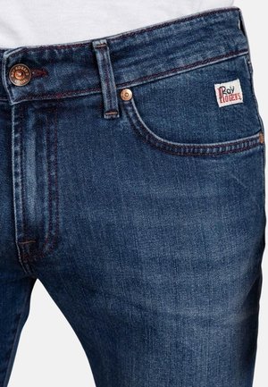 Jeans in denim di colore blu scuro con una texture liscia, dotati di chiusura a bottone metallico, cuciture visibili e una piccola etichetta del marchio sulla tasca.