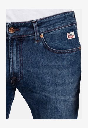 Jeans in denim di colore blu scuro con una texture liscia, dotati di chiusura a bottone metallico, cuciture visibili e una piccola etichetta del marchio sulla tasca.