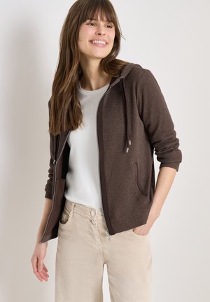 Uśmiechnięta kobieta w brązowej bluzie na zamek, nałożonej na białą koszulę, oraz w beżowych spodniach, stojąca z jedną ręką w kieszeni na neutralnym tle.