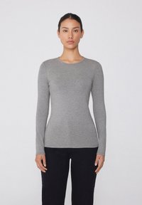 Langärmliges, enganliegendes graues Oberteil aus glattem, weichem Stoff mit rundem Ausschnitt und minimalen Nähten. Kombiniert mit schwarzen Hosen.
