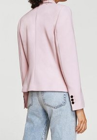 Blazer rose clair sur mesure, au design ajusté, texture lisse et boutons décoratifs sur les manches, associé à un jean en denim clair.