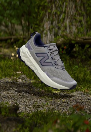 Szary i granatowy but do biegania New Balance unoszący się nad leśną podłożem z żwirem i trawą, z drzewami pokrytymi mchem w tle.