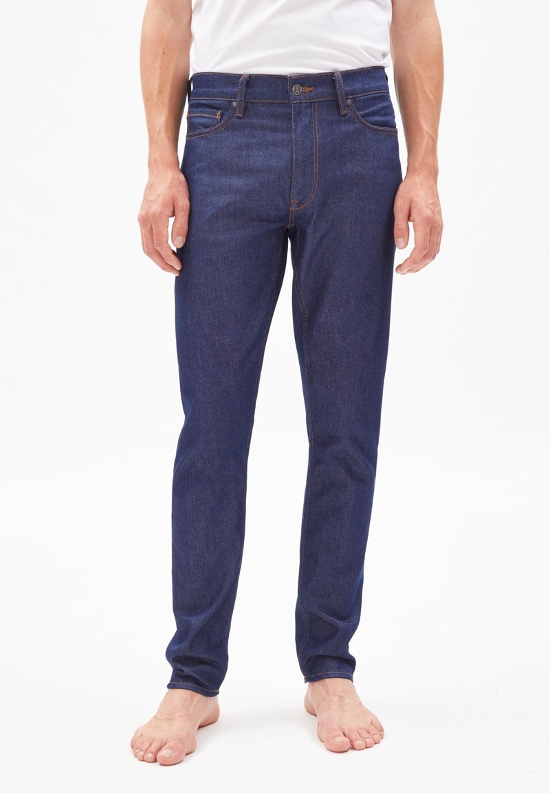 Jeans en denim bleu foncé avec une coupe slim, une couture solide, un design à cinq poches et une ceinture ornée d'une étiquette de marque.