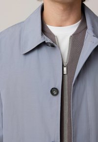 Jachetă gri deschis cu guler ascuțit, un singur buton negru și textură netedă. Sub aceasta, un strat gri cu fermoar are accente cu nervuri.