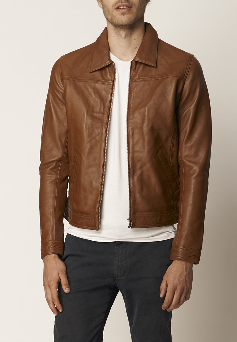 Braune Lederjacke mit einem Frontreißverschluss, breitem Kragen und subtilen Nähten; kombiniert mit einem weißen T-Shirt und dunklen Hosen.