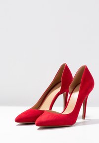 Talons aiguilles en daim rouge avec un bout pointu et des côtés échancrés, présentant une doublure intérieure lisse et un talon stiletto fin.