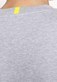 Tessuto di cotone grigio con texture a coste, caratterizzato da un'etichetta gialla a quell'altezza sul retro del collo. Design semplice e minimal.