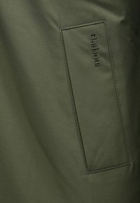 Detalhe de uma jaqueta verde-oliva com um bolso costurado e o logótipo "Barbour" em relevo. Tecido liso e mate com textura subtil.