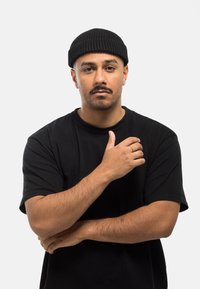Schwarze gerippte Mütze und übergroßes schwarzes T-Shirt, mit einem goldenen Ring an der rechten Hand, der auf dem linken Arm ruht, vor einem weißen Hintergrund.